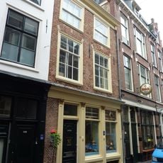 Pand met gepleisterde gevel onder rechte houten kroonlijst, met t-vensters en lage 6-ruitsschuifvensters meervoudige omlijstingen, in twee verdiepingen, afgedekt door zadeldak, evenwijd