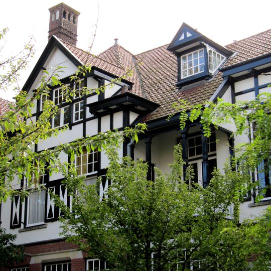 Linkerdeel van een vrijstaand bouwblok bestaande uit twee symmetrische villa's, gebouwd in overgangsarchitectuur, gecombineerd met kenmerken van de 'Stick-style'