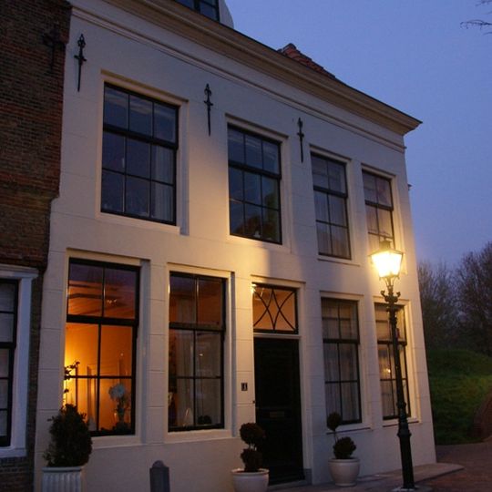 Gepleisterde lijstgevel voor huis van parterre en verdieping, waarop schilddak met dakvenster met vleugelstukken. Gestoken deurkalf, muurijzers en stoep