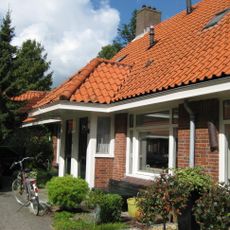 Blok met 8 woningen, gecombineerde toegangen van twee woningen onder een ingestoken schilddak, zijkant met houten topgevel, aan zuidkant toegangspoort tot binnenterrein.