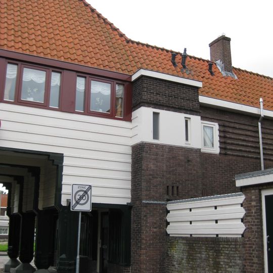 Poortgebouw met twee winkels met bovenwoningen, met veel hout zoals de zware kolommen in poort, bouwstijl Amsterdamse School, ligging aan Purmerweg centraal deel van Tuindorp Nieuwendam