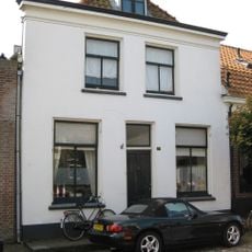 Pand met eenvoudige witgepleisterde lijstgevel. Beneden vensters met luiken