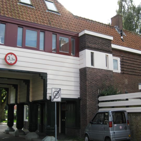 Poortgebouw met twee winkels met bovenwoningen, met veel hout zoals de zware kolommen in poort, bouwstijl Amsterdamse School, ligging aan Purmerweg centraal deel van Tuindorp Nieuwendam