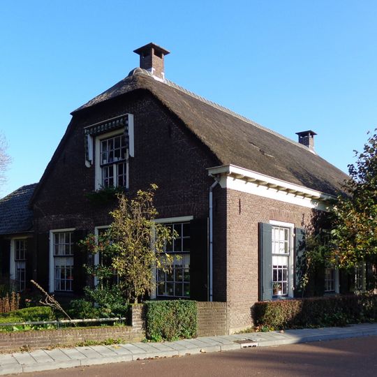 Lage woning onder rieten zadeldak aan de korte zijden afgewolfd. Kleine roeden, schuiframen, muurankers