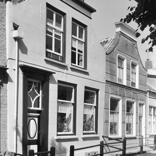 Huis met gepleisterde lijstgevel met ingezwenkte top. Schuifvensters. Voordeur met na-empire panelen en een omlijsting uit dezelfde periode