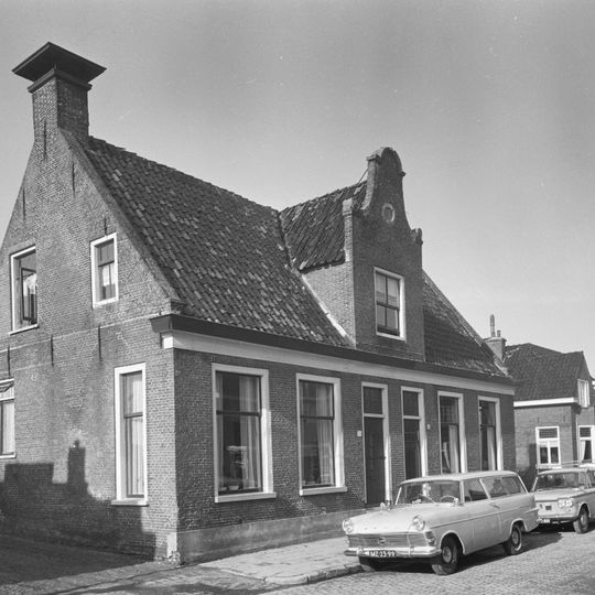 Groot dorpshuis met ingang met Vlaamse top onder zadeldak tussen topgevels waarop hoekschoorstenen met borden