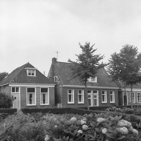 Pand onder hoge dwarskap tegen zijgevel van gele steen met rode vlechtingen, onder een dak met nr. 99