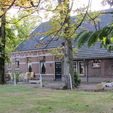 De Mans: boerderij van het hallehuistype opgetrokken in bruine baksteen onder een met zwarte muldenpannen gedekt zadeldak dat boven het uitgebouwde woonhuisgedeelte is geknikt