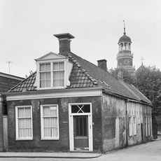 Eenvoudig pandje onder voor afgeschuind zadeldak met grote dakkapel en topschoorsteen waarop bord