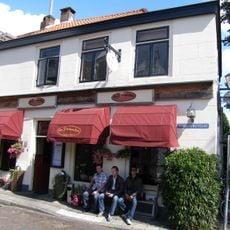 Gedeeltelijk verhoogd hoekpand met gepleisterde rechte gevels, diep doorlopend langs het grachtje