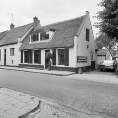 Gepleisterd dwarshuis onder rieten zadeldak tussen puntgevels. In de voorgevel vensters met luiken en negenruitsschuiframen