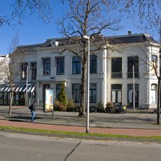 Twee bouwlagen tellend blokvormig witgepleisterd pand onder met platte friese pannen gedekt afgeknot schilddak