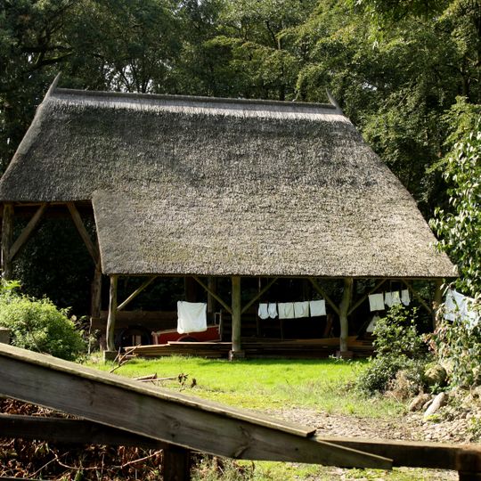 Boerderij, houten kapschuur. Terzijde van de boerderij staat een open houten kapschuur, riet gedekt en met een sporenkap ter lengte van vier vakken.