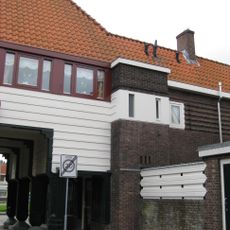 Poortgebouw met twee winkels met bovenwoningen, met veel hout zoals de zware kolommen in poort, bouwstijl Amsterdamse School, ligging aan Purmerweg centraal deel van Tuindorp Nieuwendam