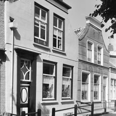 Huis met gepleisterde lijstgevel met ingezwenkte top. Voordeur en vensteromlijstingen met afgeschuinde hoeken. Voordeur met na-empire velden.