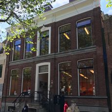 Vijf traveeën breed pand met vernieuwde gevel met middenrisaliet, hoekpilaster en rechte kroonlijst onder schilddak met twee hoekschoorstenen met borden