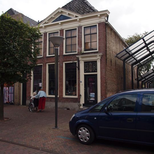 Vier traveeën breed pand met omlijste ingang en rechte kroonlijst met timpaan onder voor afgeschuind schilddak met topschoorsteen