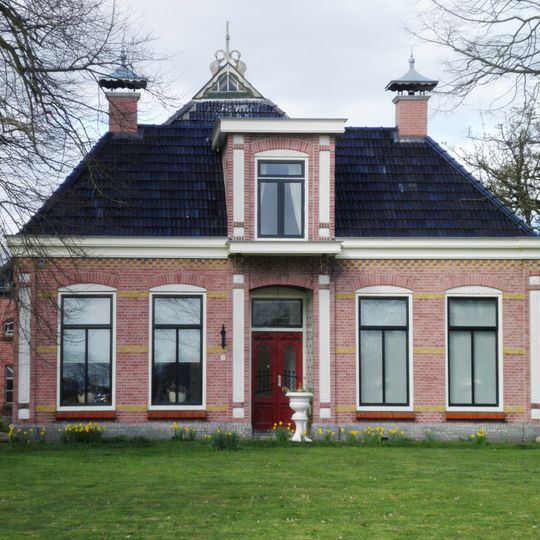 Dwarshuisboerderij in roze kalkzandsteen met dwarshuis onder schilddak gedekt met zwarte Friese pannen en met versierde dakkapel en twee schoorstenen op nokhoeken