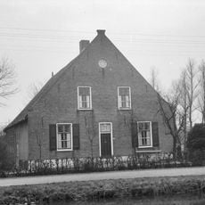 Boerderij, waarvan het woongedeelte een afgeknotte puntgevel met sierankers heeft en links een opkamer. Hogere houten stal onder een rieten dak met het woongedeelte