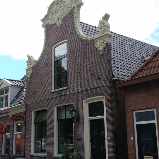 Woonhuis onder zadeldak tegen klokgevel met gesneden deur en bovenlicht, zwierige aanzetkrullen en groot kuifstuk in de plaatselijke Friese trant van de 18e eeuw