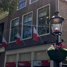 Hoog pand tegen klokgevel met natuurstenen aanzetkrullen en kroonlijst met emblemen onder zadeldak