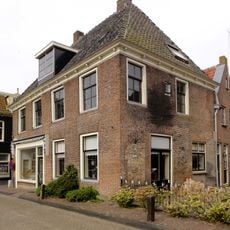 Aan drie zijden vrijstaand breed pand met jongere pui en smalle geprofileerde rondgaande daklijst met blokjes onder fors schilddak met jongere dakkapel