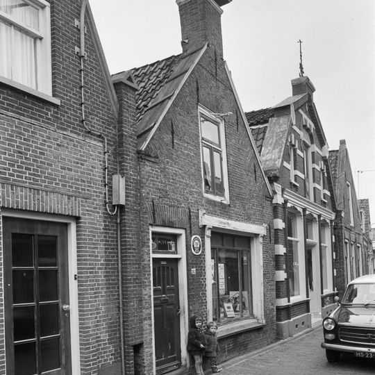 Pand onder zadeldak tegen topgevel met vlechtingen langs de zijden op aanzetlijsten en forse topschoorsteen waarop bord