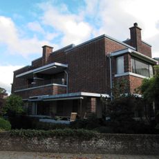 Twee geschakelde herenhuizen die onder een hoek van 45 graden ten opzichte van de rechthoekige kruising liggen, bestaande uit twee bouwlagen onder platte daken