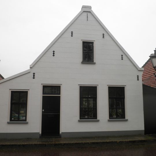 Woning met gepleisterde puntgevel onder zadeldak waarvan het linker schild dieper doorloopt dan het rechter