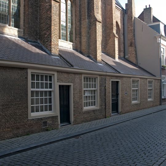 Tegen het koor der kerk gebouwd ondiep pand met lijstgevel waarin ramen met kleine roedenverdeling