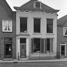 Huis onder schilddak en met empire lijstgevel, gepleisterd, waarin door pilasters omlijste ingang, kroonlijst op voluutvormige consoles en versierd met guirlandes