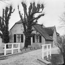 Boerderij onder rieten wolfdak met rechts een uitgebouwde zijkamer met puntgevel. In de gepleisterde voorgevel vensters met luiken en zesruitsschuiframen