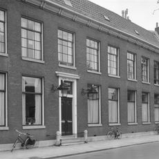 Pand met statige gevel met rechte kroonlijst, ter weerszijden van de ingang fraaie lantaarns