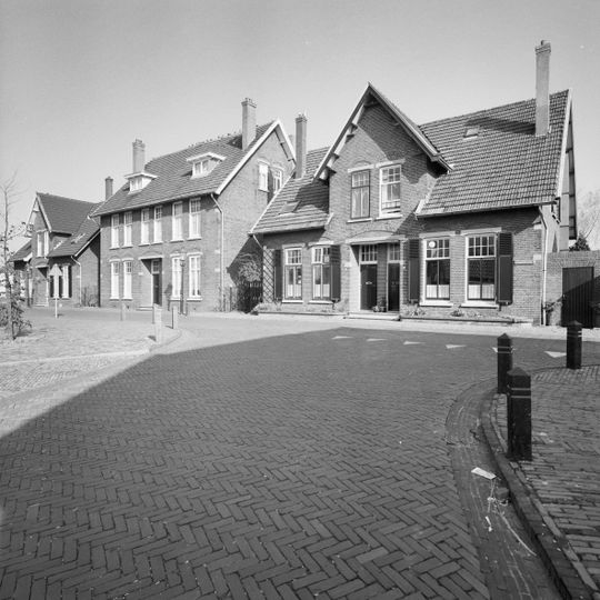 Dubbel woonhuis gebouwd op een T-vormige plattegrond en bestaande uit een eenlaags hoofdbouwmassa onder een overstekend zadeldak gedekt met gesmoorde en rode muldenpannen